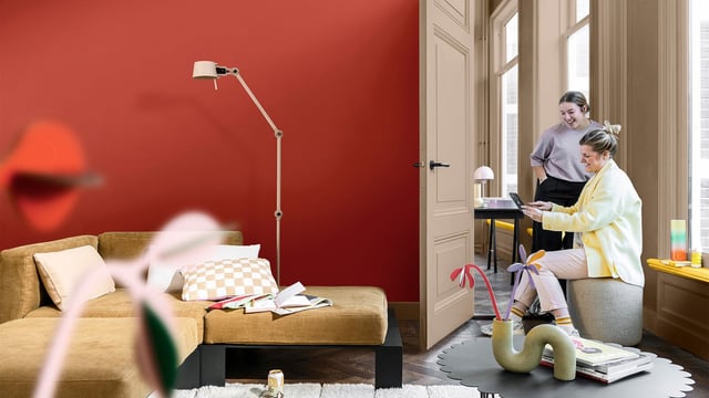 Bright living room with a red wall and sofa; two people unpacking near the doorway.