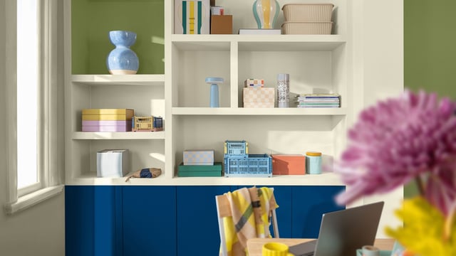Bright home office with blue cabinets, white shelves and a green wall; blurred purple flower in the foreground.