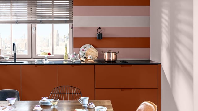 Warm kitchen interior with terracotta cabinets, a striped wall, and a table with cups and a teapot.