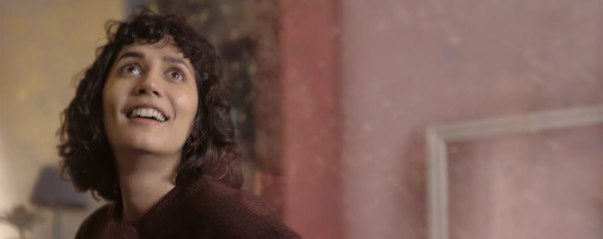 Smiling woman with curly hair looking up indoors by a painted wall and open doorway.