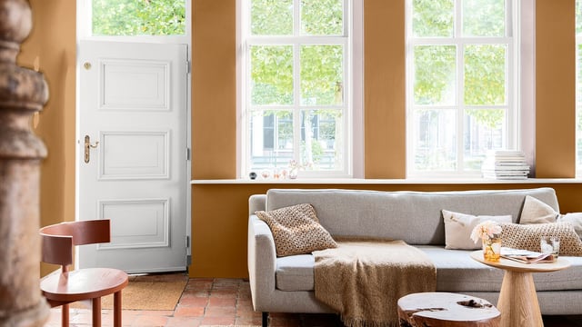 Bright living room with a beige sofa by large windows, a white door and a table lamp on a side table.
