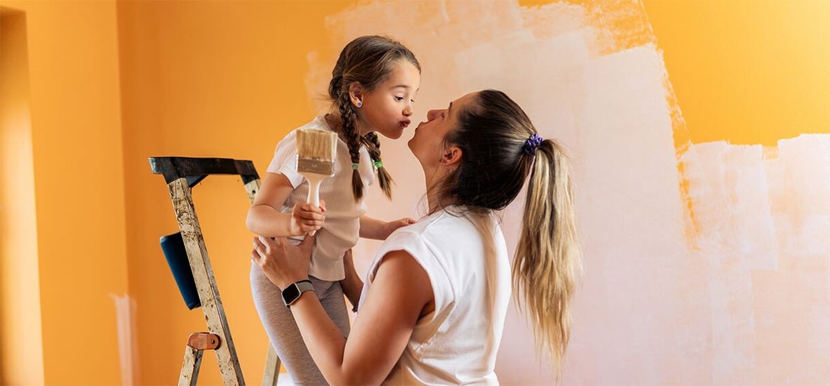 Woman helps a young girl on a ladder as they paint an orange wall white, sharing a nose-to-nose moment.