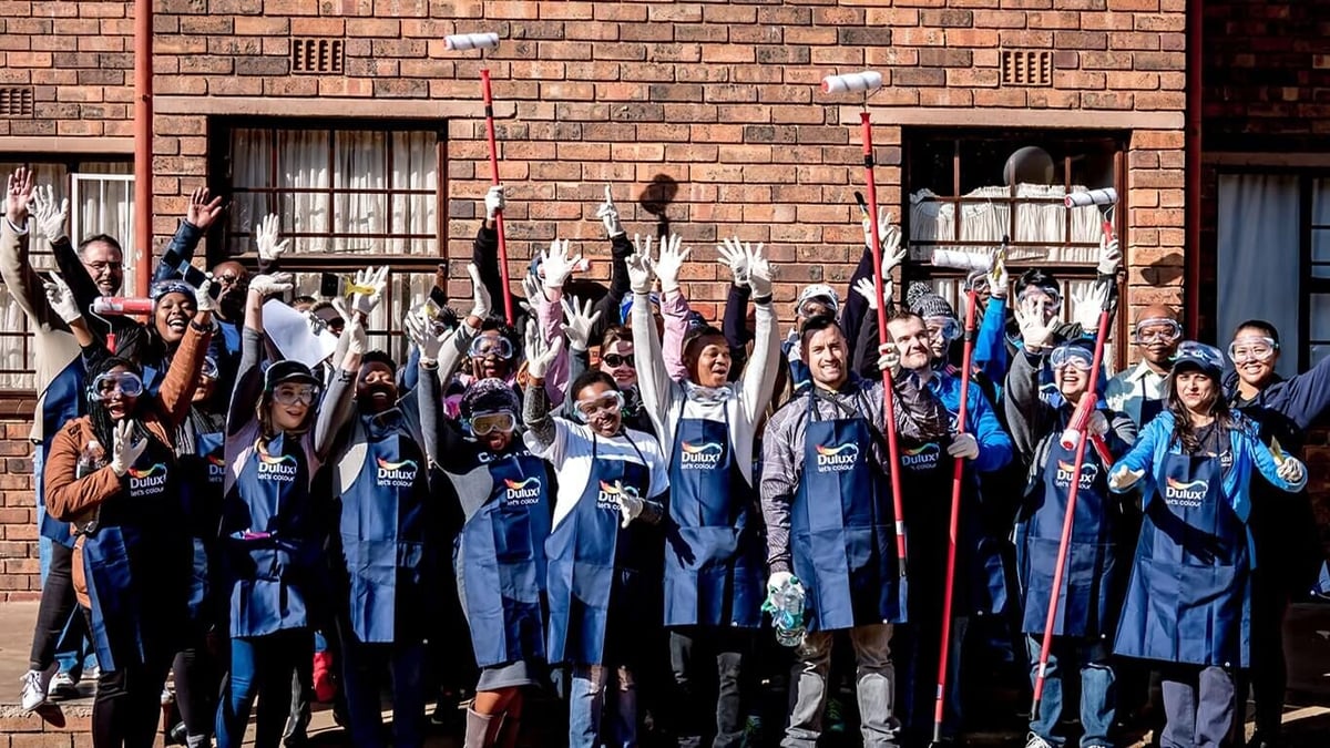 Group of children in blue Dulux aprons, smiling and waving from in front of a brick building.