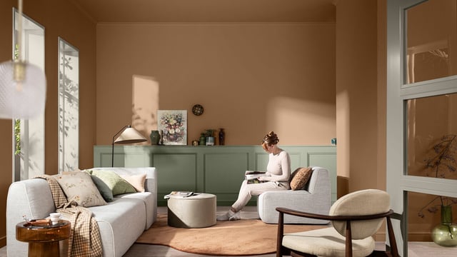 Warm, modern living room with a sofa, coffee table, plants, and a person reading by the window.