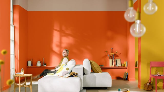 Modern living room with an orange wall, a person on the sofa, and colorful decor in a warm interior.