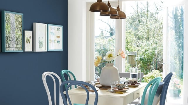 Bright dining area with table by large window, pendant lights, and blue chairs against a blue wall.
