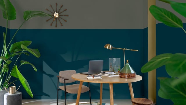 Modern home office corner with round wooden desk, laptop, plants, and a teal accent wall.