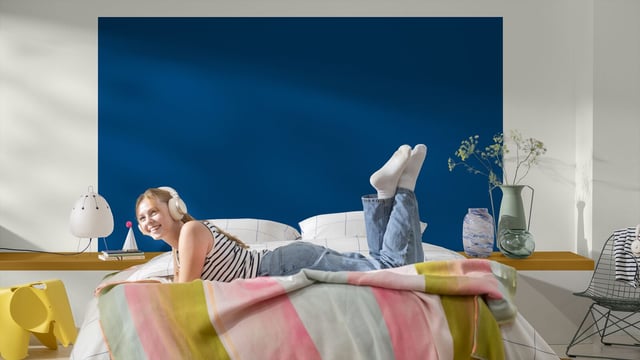 Woman lounging on a bed in a modern bedroom with a colorful blanket, blue accent wall and a vase of flowers